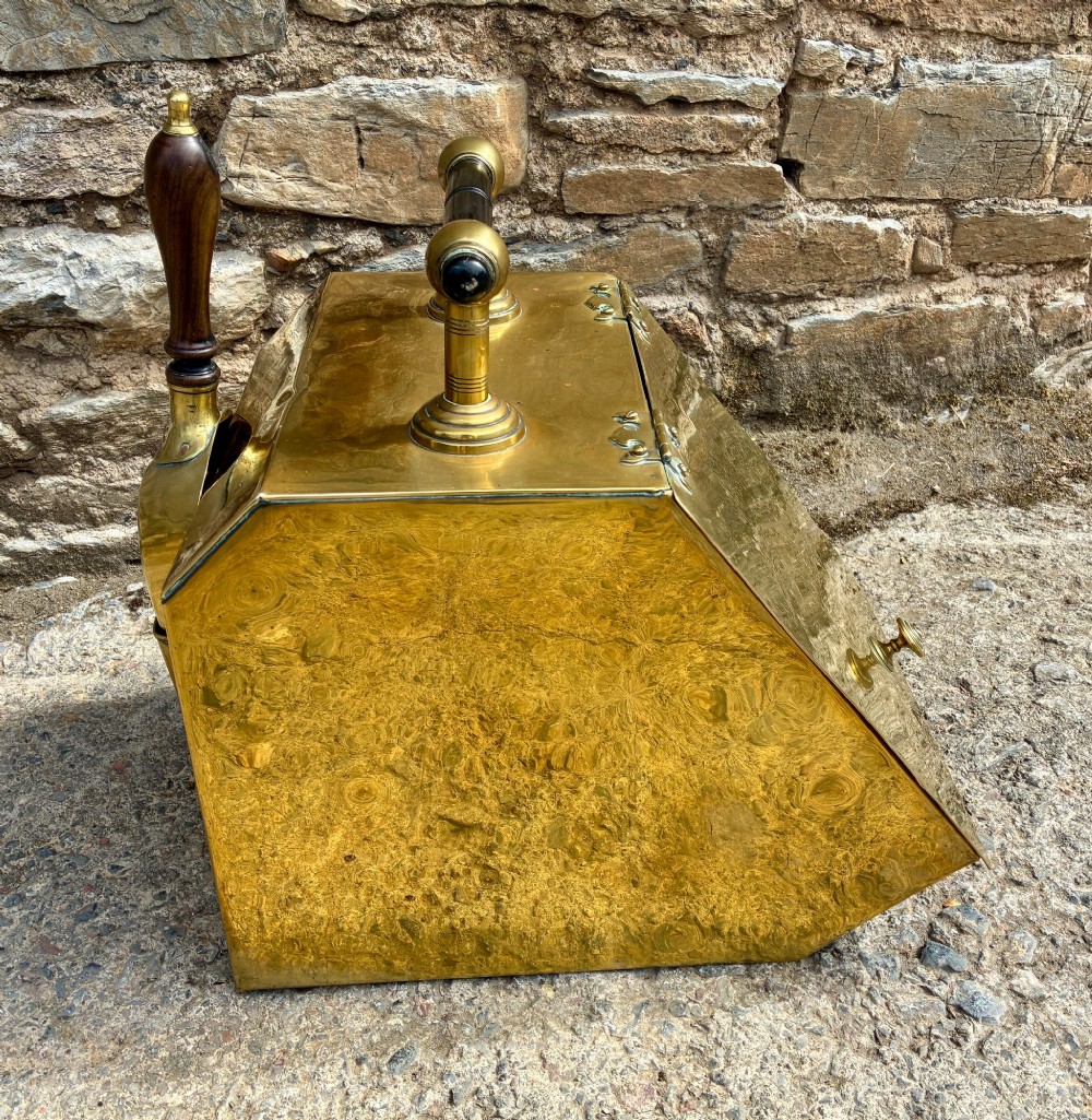 An antique brass sloped coal scuttle featuring a turned wooden carrying handle, a hinged lid, and an attached brass shovel at the rear. View V