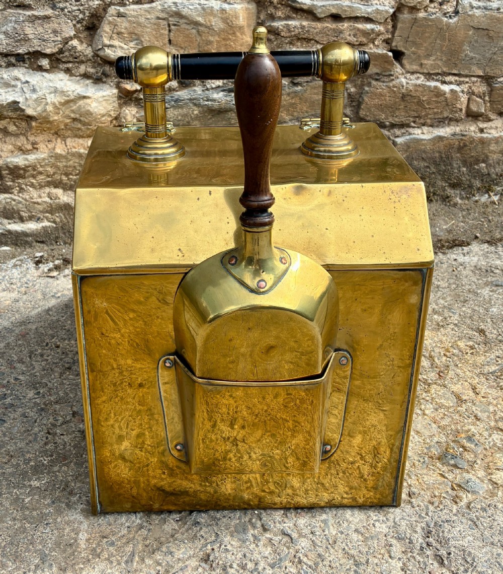 An antique brass sloped coal scuttle featuring a turned wooden carrying handle, a hinged lid, and an attached brass shovel at the rear. View VI