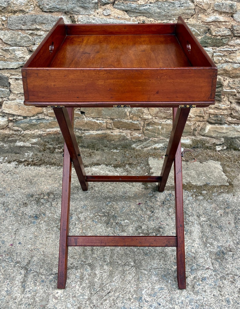 A Victorian mahogany butlers tray with cut-out hand holes resting on a folding wooden X-frame stand. View V