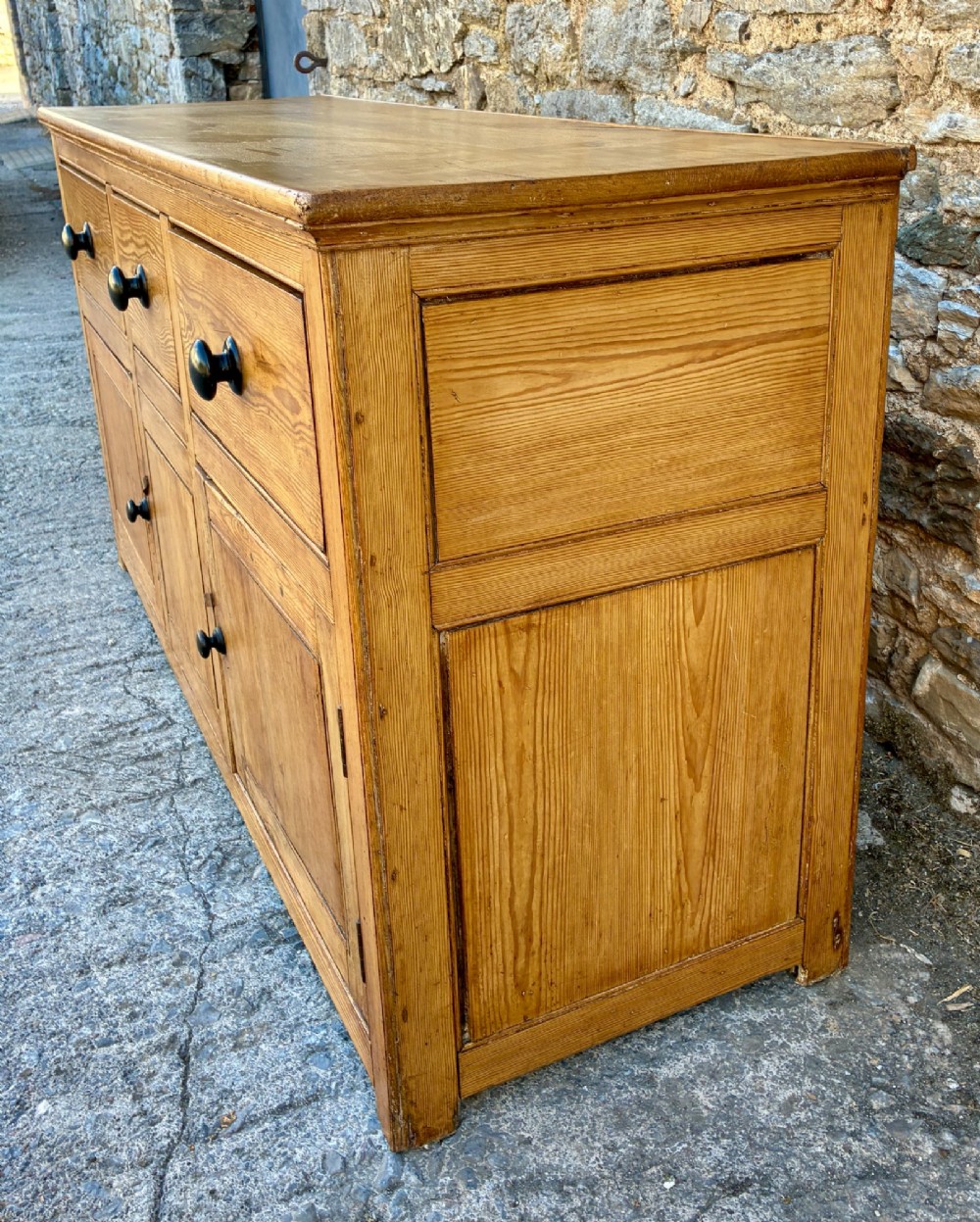 A nineteenth-century pine dresser base featuring three top drawers, three lower panelled cupboard doors, and turned dark wooden knobs. View VII