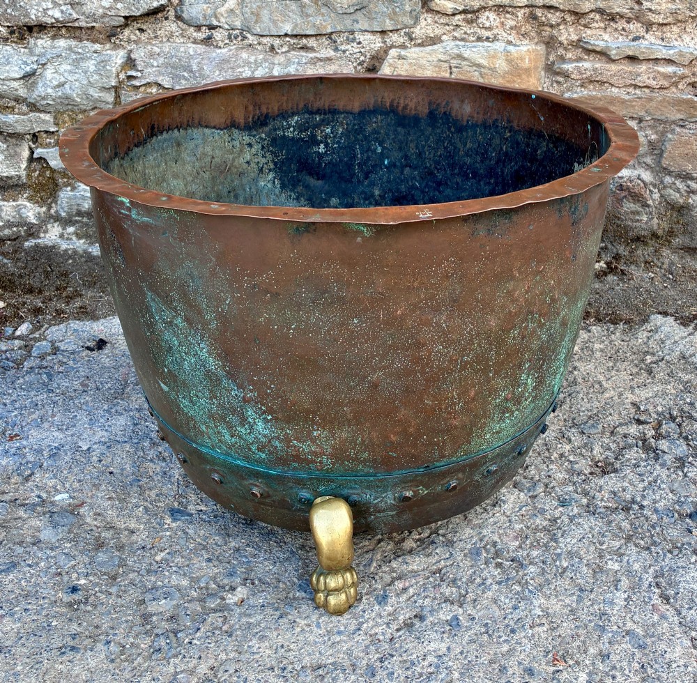 A large round copper boiler with a hand-riveted base seam and flared rim, supported on four decorative cast brass claw feet. View V