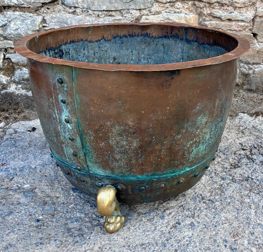 A large round copper boiler with a hand-riveted base seam and flared rim, supported on four decorative cast brass claw feet. View VII