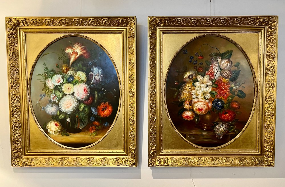 A pair of oil paintings on board featuring floral bouquets framed within oval apertures inside ornate gold frames.