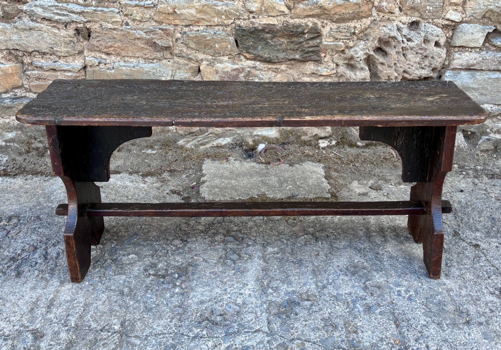 A long antique dark wooden hall bench featuring a flat plank seat, shaped slab end supports, and a lower wooden stretcher.
