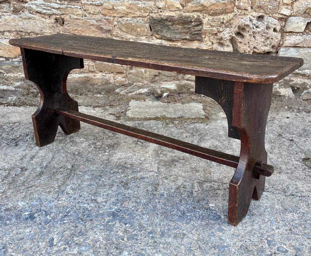 A long antique dark wooden hall bench featuring a flat plank seat, shaped slab end supports, and a lower wooden stretcher. View IV