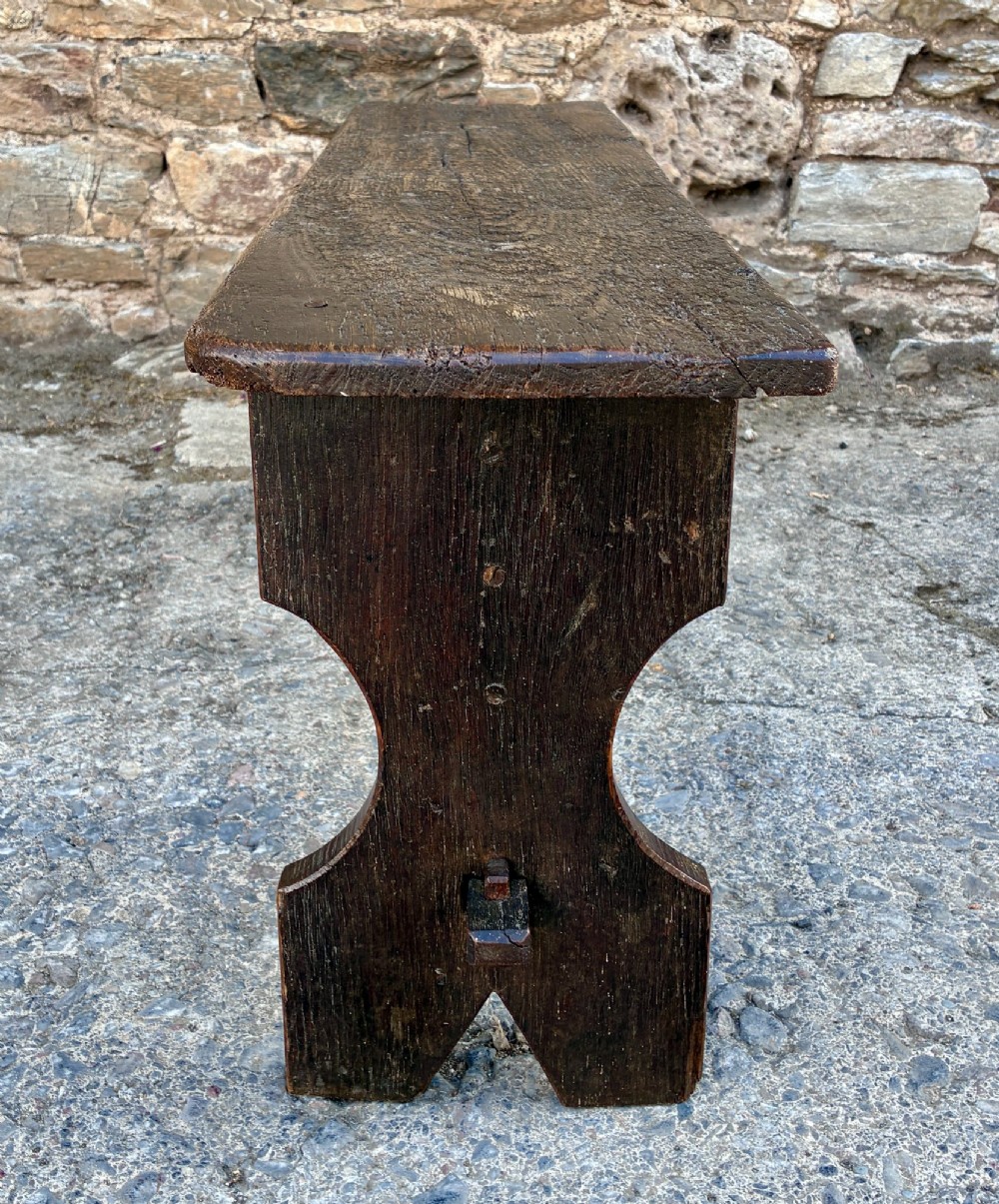 A long antique dark wooden hall bench featuring a flat plank seat, shaped slab end supports, and a lower wooden stretcher. View V