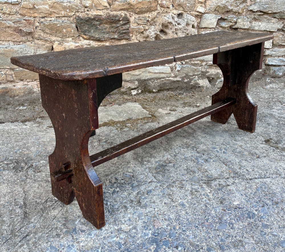 A long antique dark wooden hall bench featuring a flat plank seat, shaped slab end supports, and a lower wooden stretcher. View IX