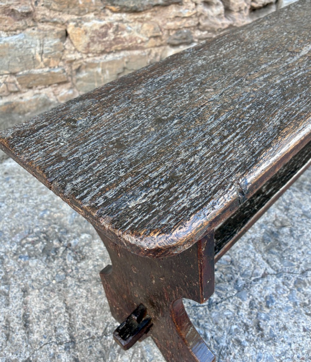 A long antique dark wooden hall bench featuring a flat plank seat, shaped slab end supports, and a lower wooden stretcher. View XI