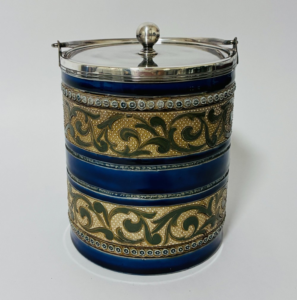 A cylindrical Victorian Doulton Lambeth stoneware biscuit barrel featuring cobalt blue and beige scrollwork decoration, fitted with a silver-plated lid, swing handle, and finial.