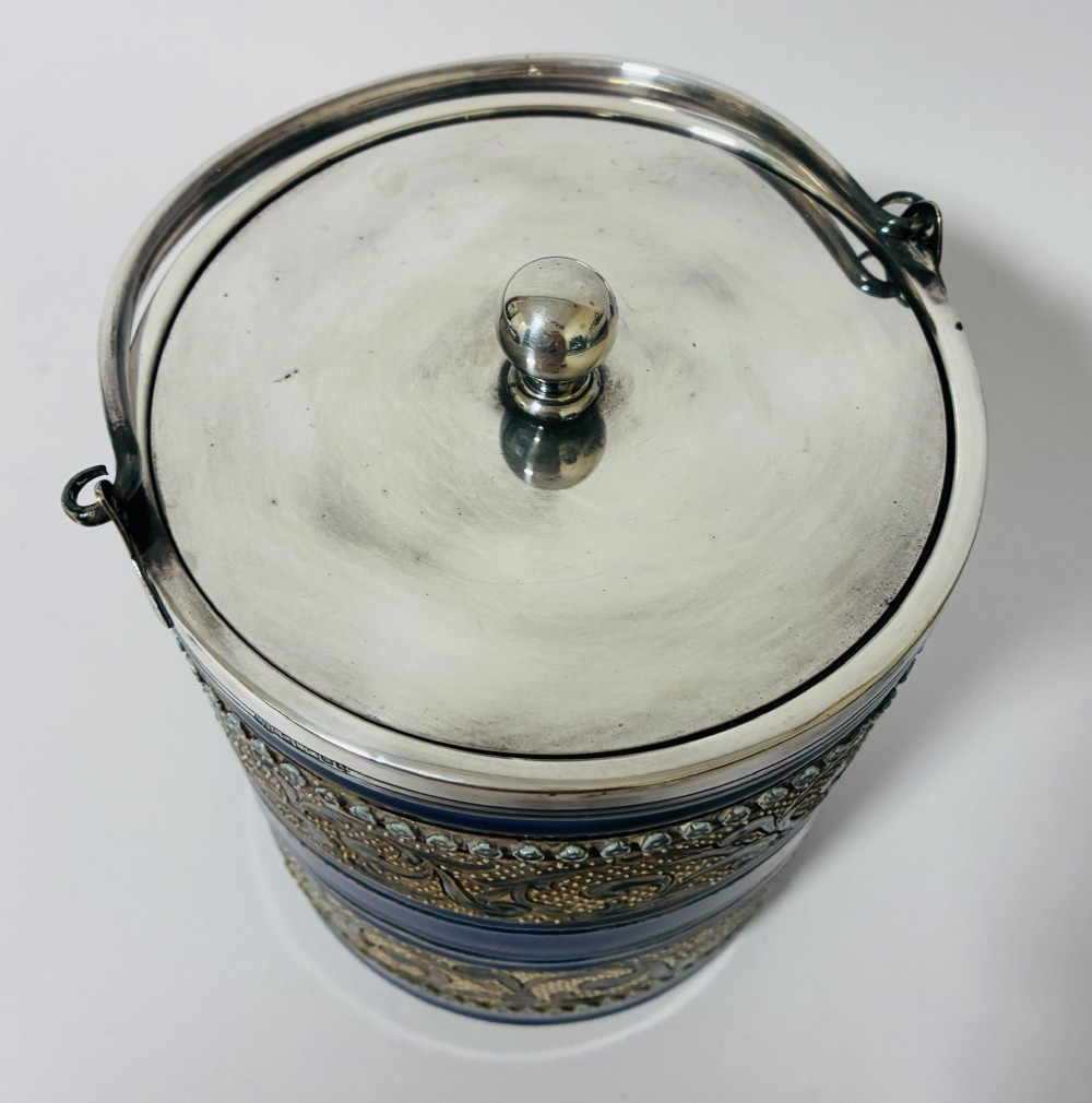 A cylindrical Victorian Doulton Lambeth stoneware biscuit barrel featuring cobalt blue and beige scrollwork decoration, fitted with a silver-plated lid, swing handle, and finial. View V
