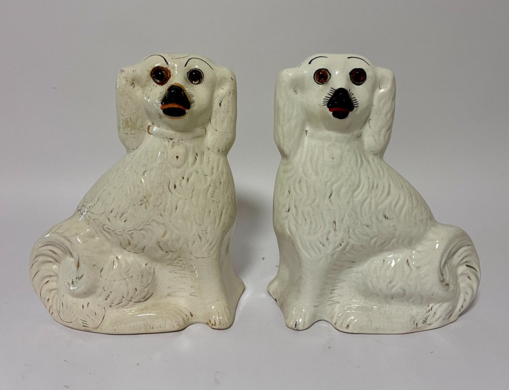 A pair of cream glazed ceramic Staffordshire spaniel figurines with black painted muzzles and amber eyes seated facing forward.