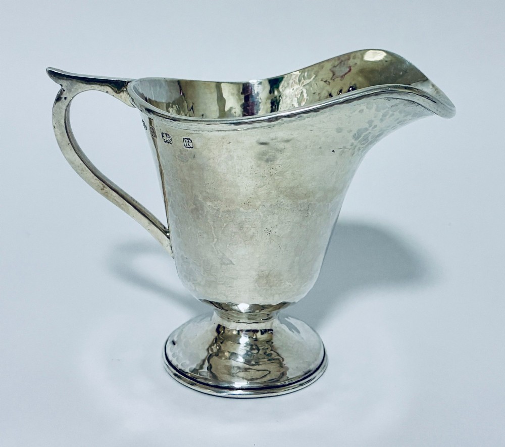 An antique sterling silver cream jug with a hand-hammered planished surface, a pedestal base, and an angular handle.