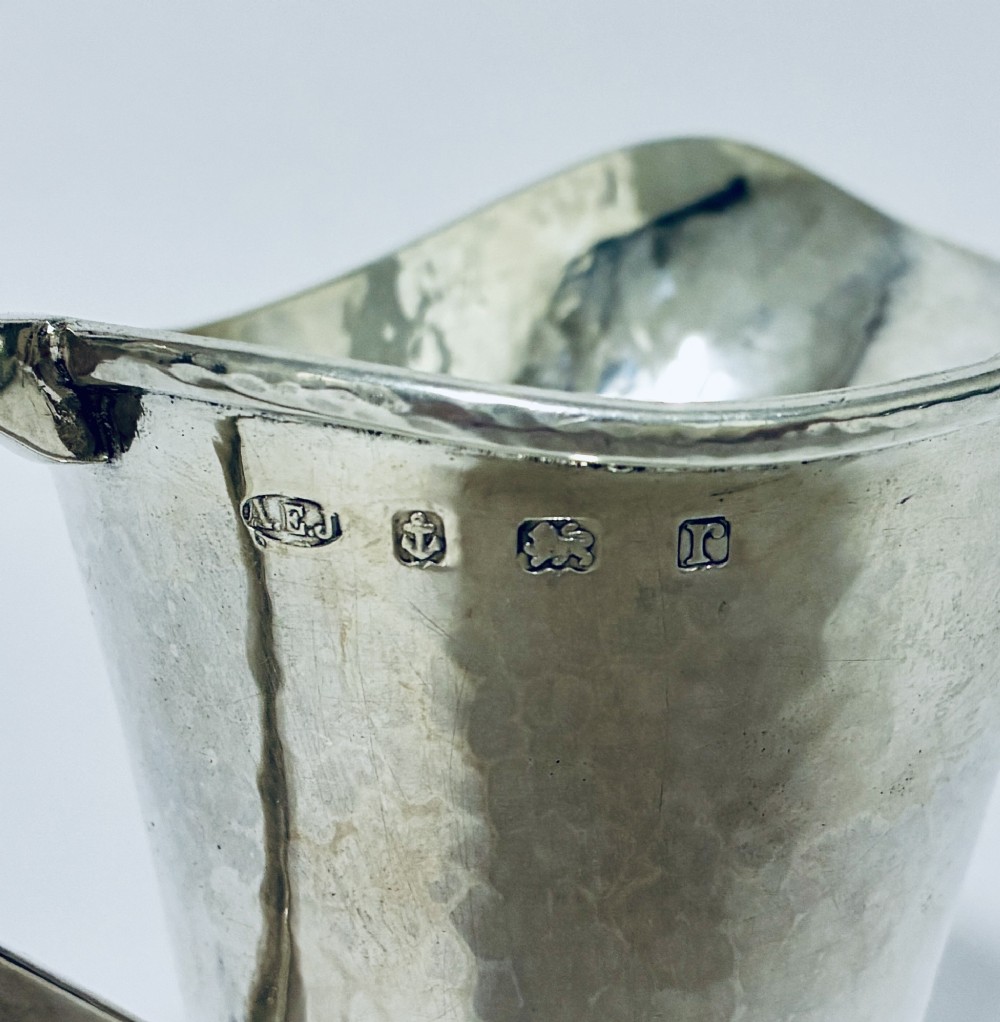 An antique sterling silver cream jug with a hand-hammered planished surface, a pedestal base, and an angular handle. View II