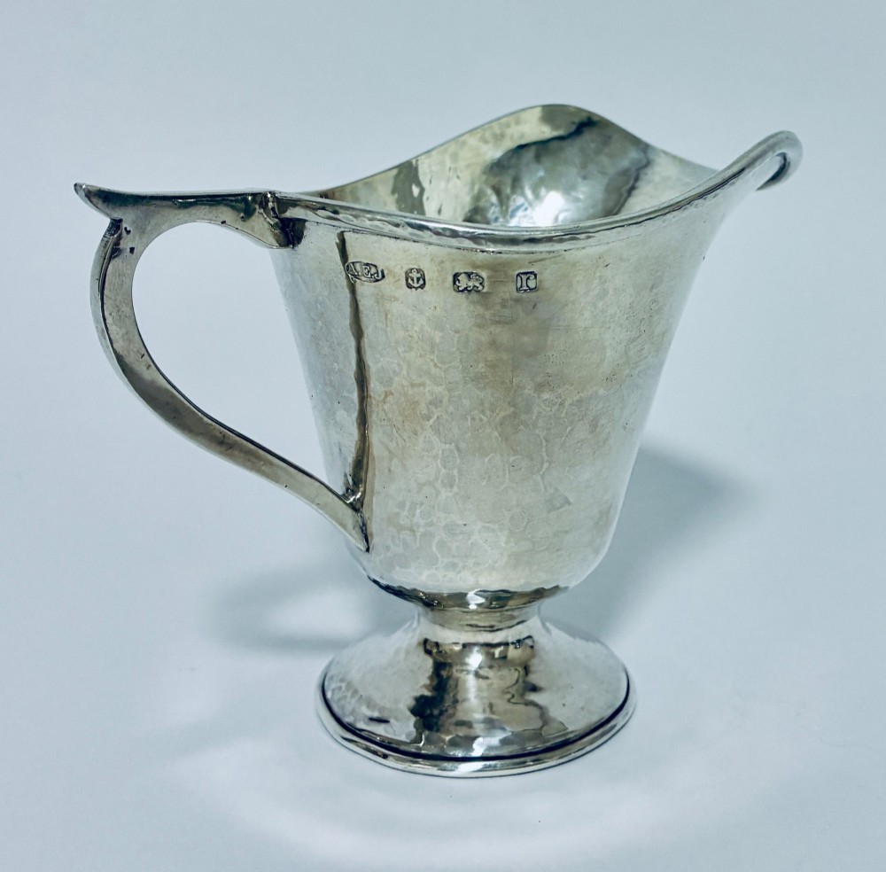 An antique sterling silver cream jug with a hand-hammered planished surface, a pedestal base, and an angular handle. View III
