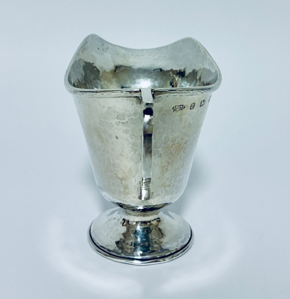 An antique sterling silver cream jug with a hand-hammered planished surface, a pedestal base, and an angular handle. View IV