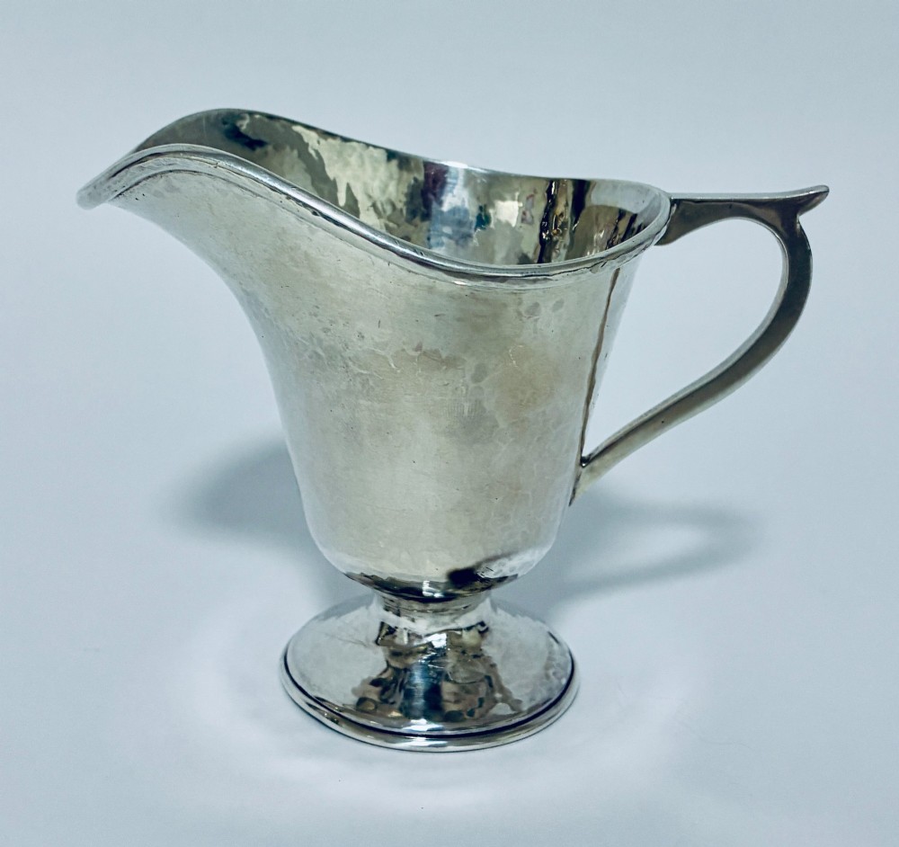 An antique sterling silver cream jug with a hand-hammered planished surface, a pedestal base, and an angular handle. View V