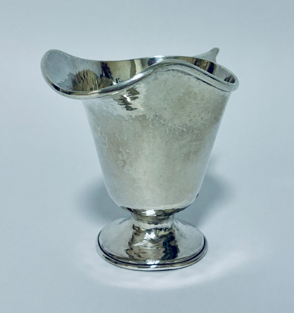 An antique sterling silver cream jug with a hand-hammered planished surface, a pedestal base, and an angular handle. View VI