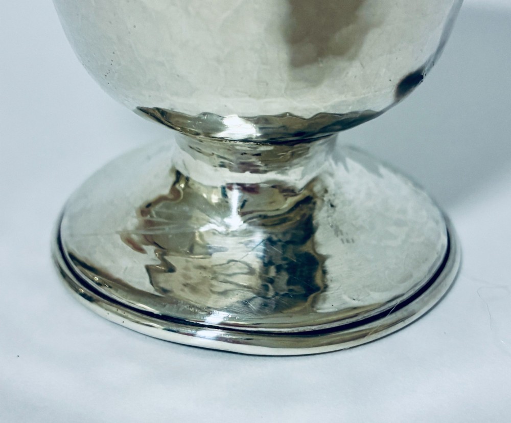 An antique sterling silver cream jug with a hand-hammered planished surface, a pedestal base, and an angular handle. View VII