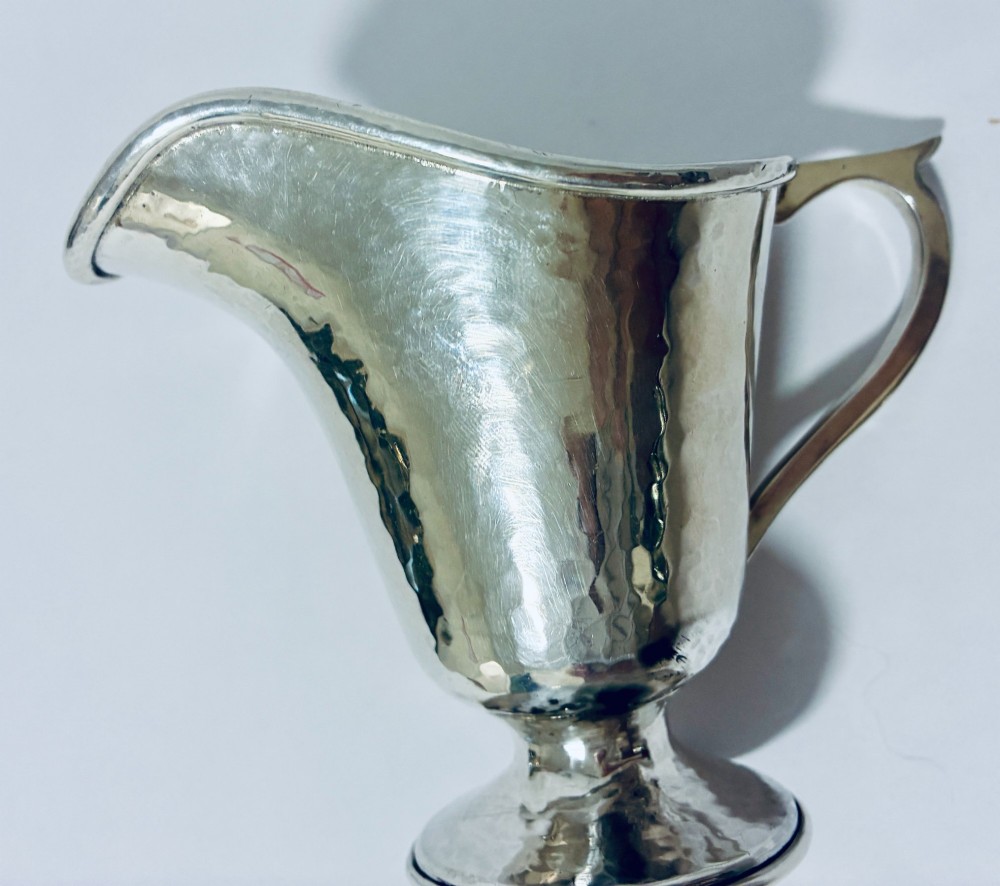 An antique sterling silver cream jug with a hand-hammered planished surface, a pedestal base, and an angular handle. View X