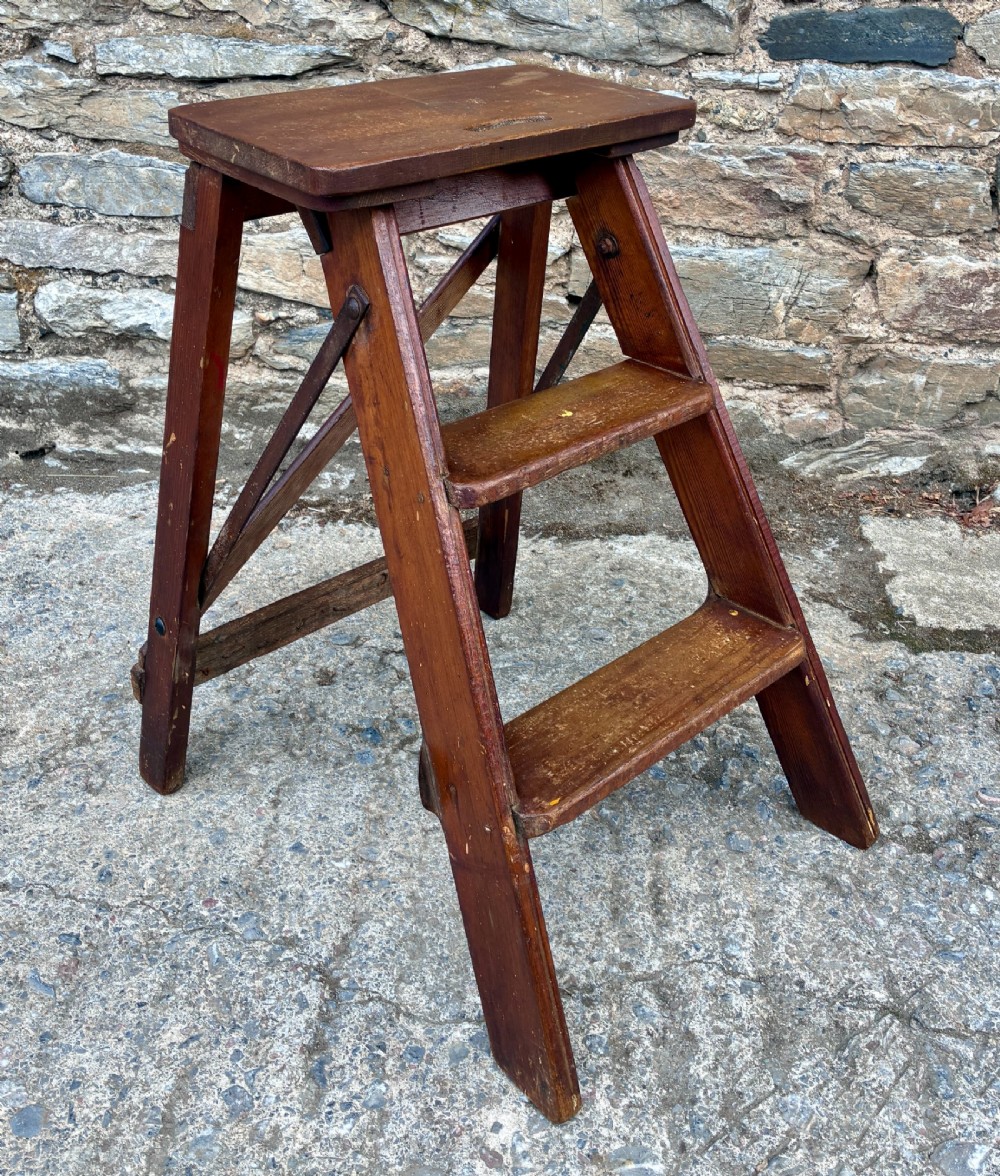 A vintage wooden folding step stool with three tread steps and metal side bracing finished in a warm reddish-brown stain.