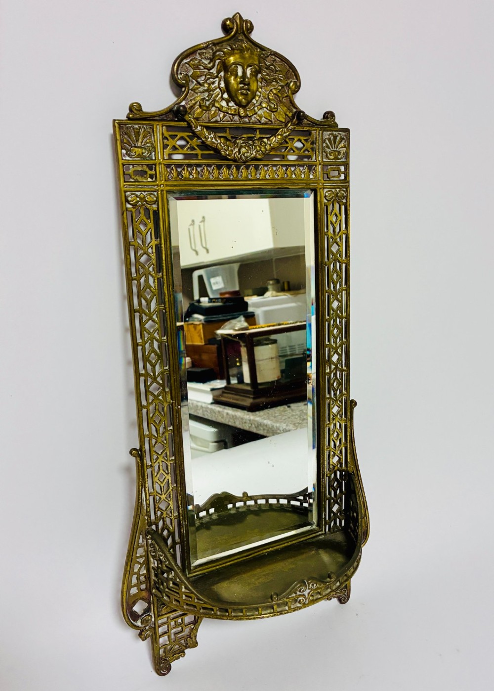 An ornate cast brass wall mirror featuring a central bevelled glass plate, openwork lattice frame, top mask cresting, and an integrated lower tray. View II