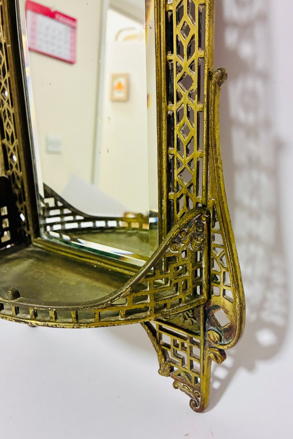 An ornate cast brass wall mirror featuring a central bevelled glass plate, openwork lattice frame, top mask cresting, and an integrated lower tray. View V