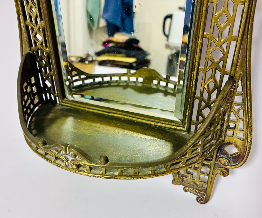 An ornate cast brass wall mirror featuring a central bevelled glass plate, openwork lattice frame, top mask cresting, and an integrated lower tray. View VI