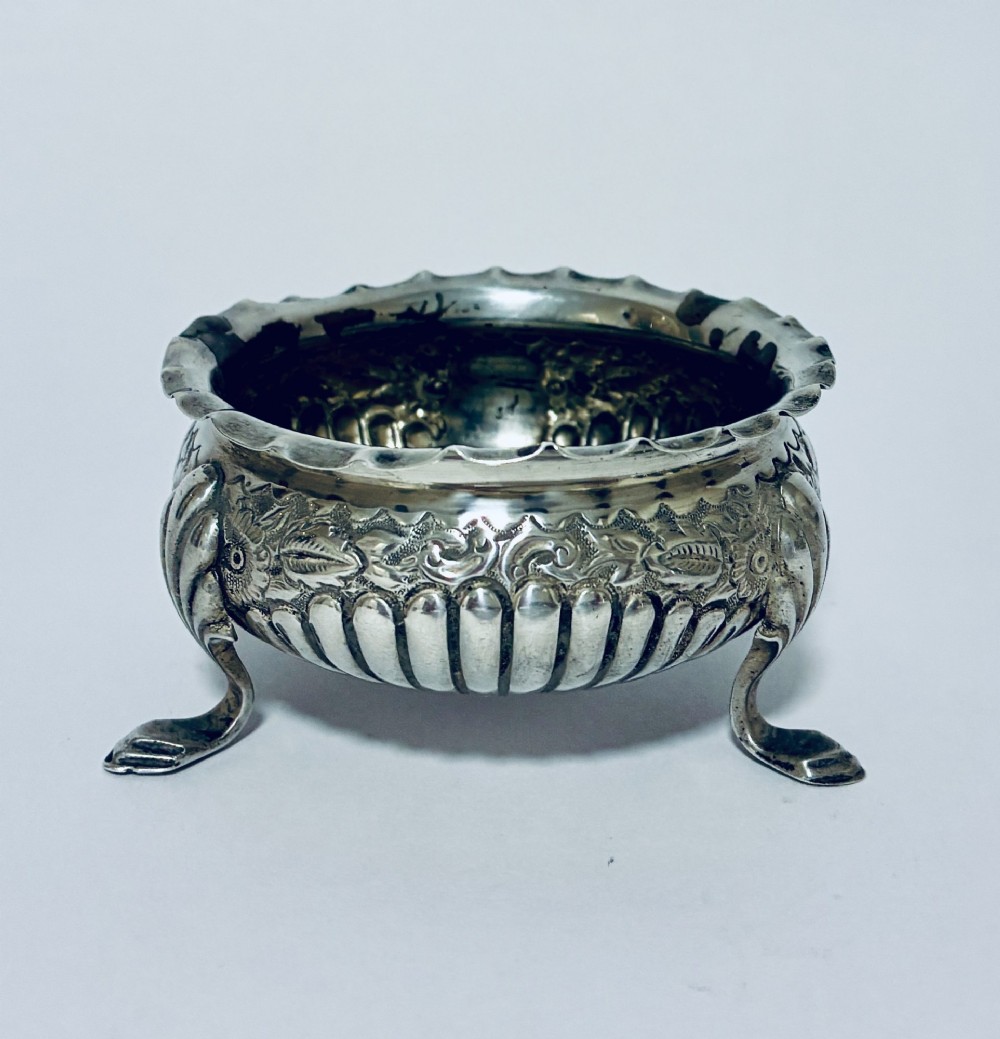 A small Victorian sterling silver salt dish with a crimped rim, chased floral decoration, fluted lower body, and three tripod legs.