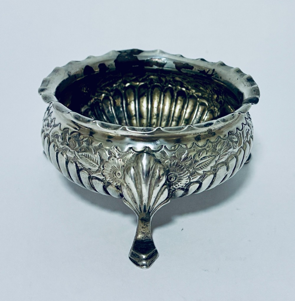 A small Victorian sterling silver salt dish with a crimped rim, chased floral decoration, fluted lower body, and three tripod legs. View II