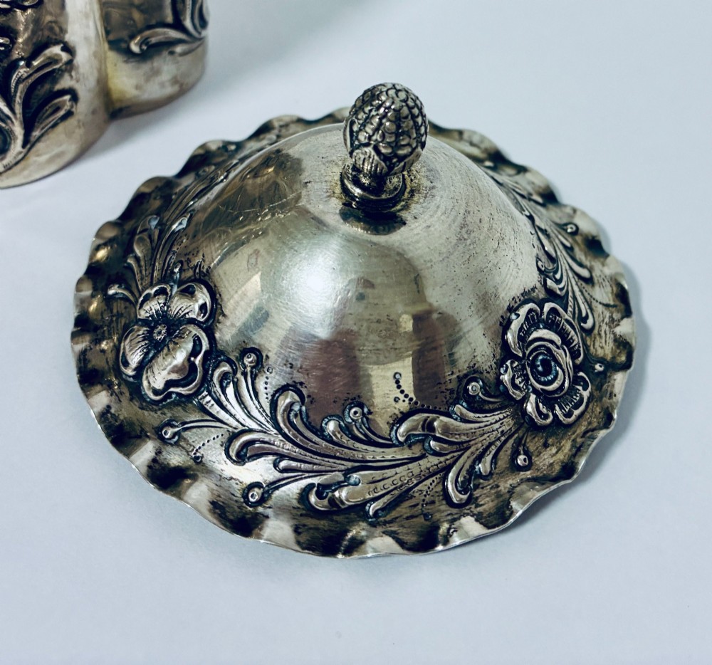 A lobed silver tea caddy with embossed floral patterns, a wavy rimmed lid, and a pinecone-shaped finial. View V