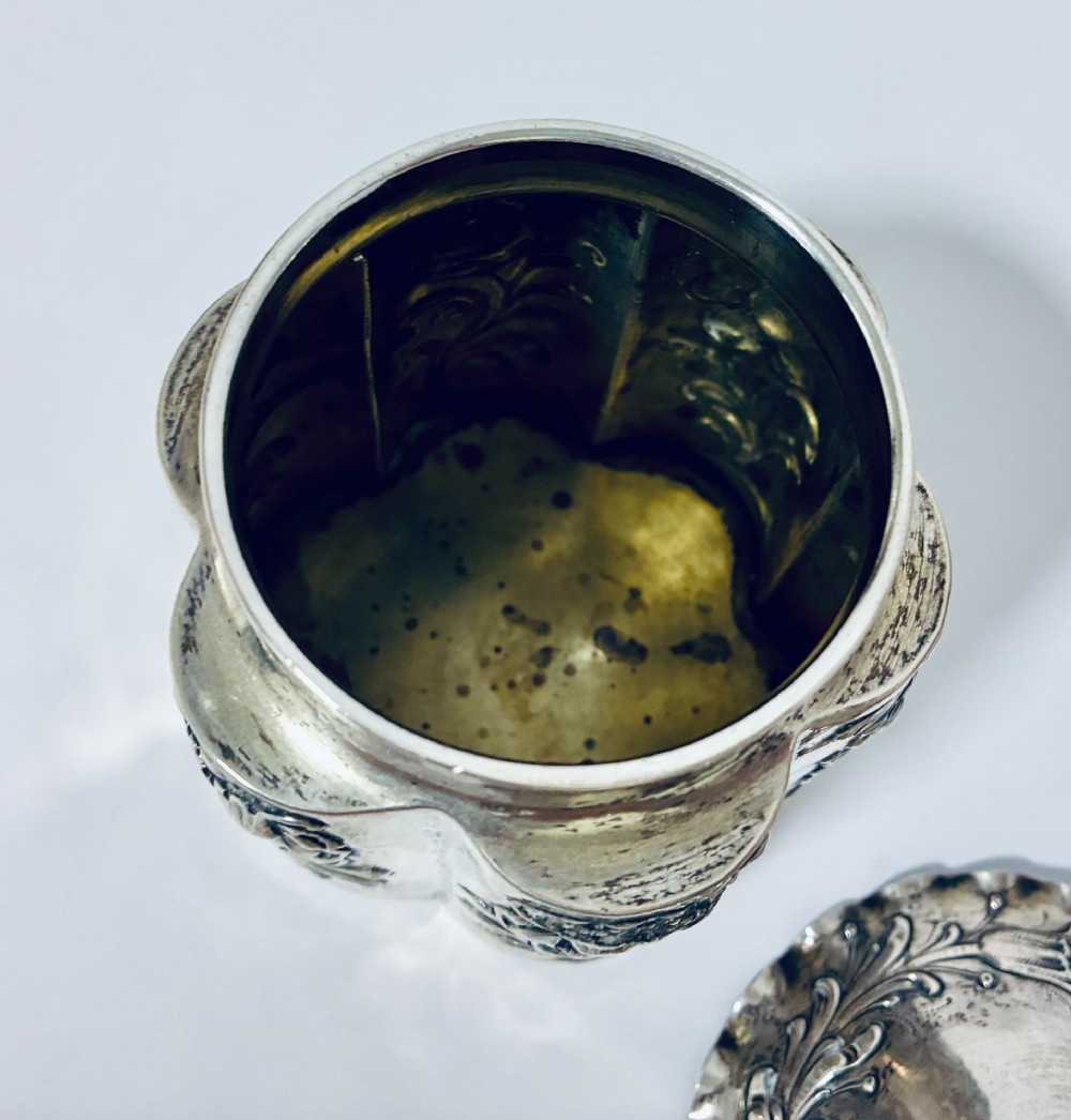 A lobed silver tea caddy with embossed floral patterns, a wavy rimmed lid, and a pinecone-shaped finial. View VI