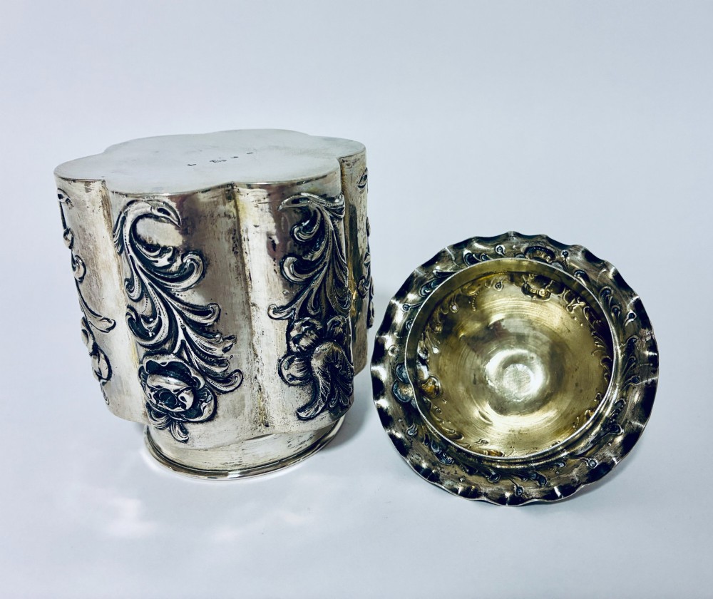 A lobed silver tea caddy with embossed floral patterns, a wavy rimmed lid, and a pinecone-shaped finial. View X