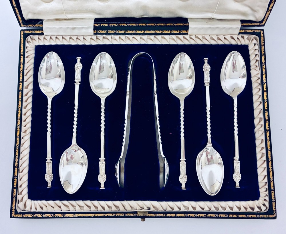 An open dark fitted presentation case lined with white satin and blue velvet containing six silver apostle teaspoons with twisted stems and matching silver sugar tongs. View II