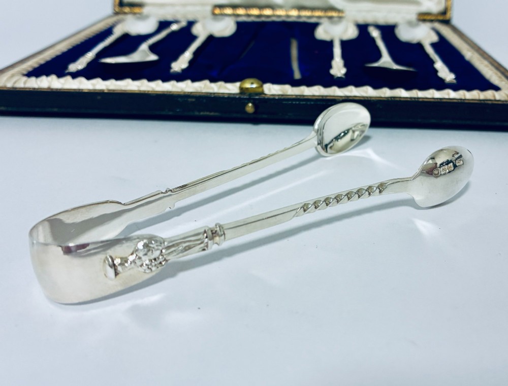 An open dark fitted presentation case lined with white satin and blue velvet containing six silver apostle teaspoons with twisted stems and matching silver sugar tongs. View V
