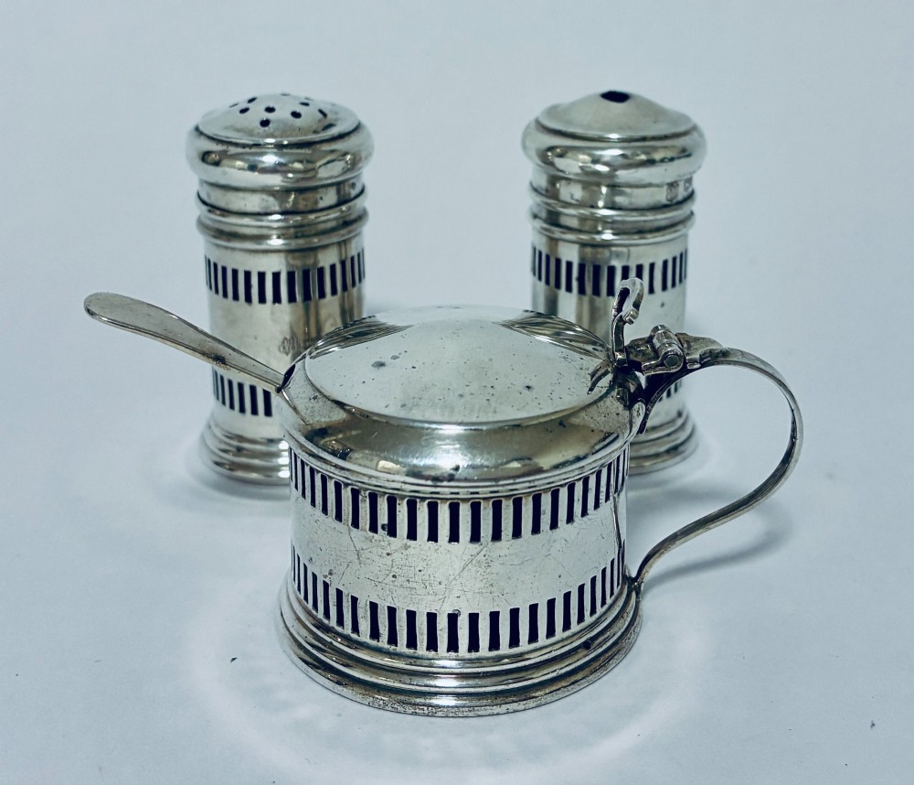 An antique three-piece silver cruet set featuring pierced vertical slot details, a mustard pot with lid and spoon, and two matching pepper casters.