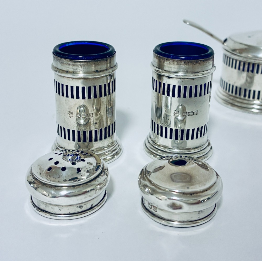 An antique three-piece silver cruet set featuring pierced vertical slot details, a mustard pot with lid and spoon, and two matching pepper casters. View VII