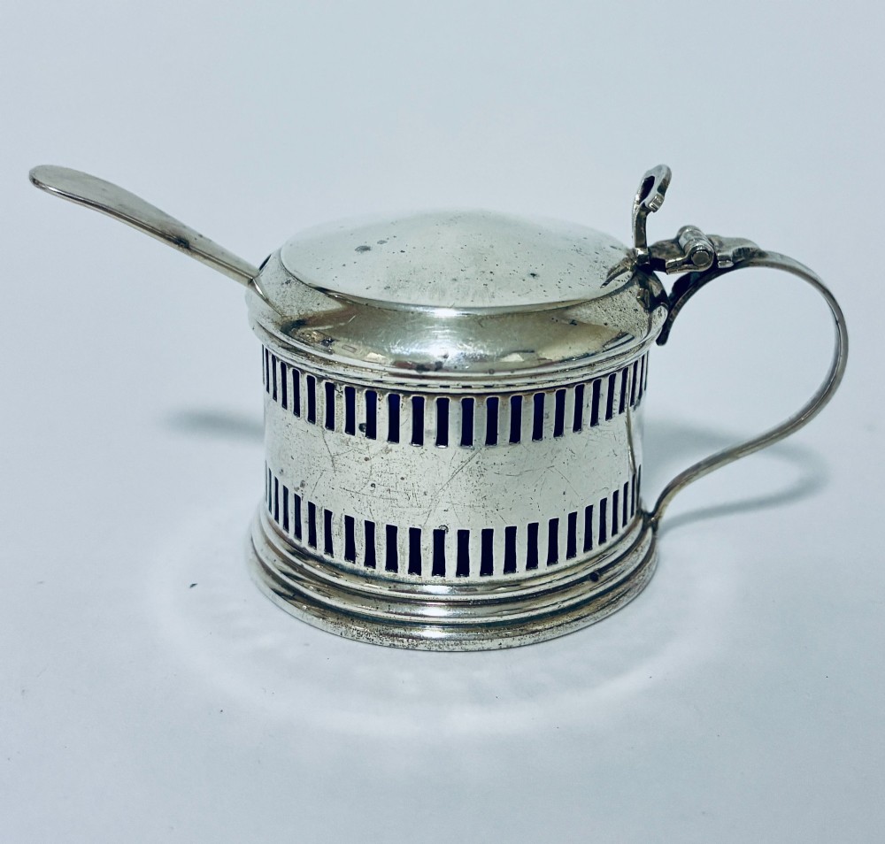 An antique three-piece silver cruet set featuring pierced vertical slot details, a mustard pot with lid and spoon, and two matching pepper casters. View X