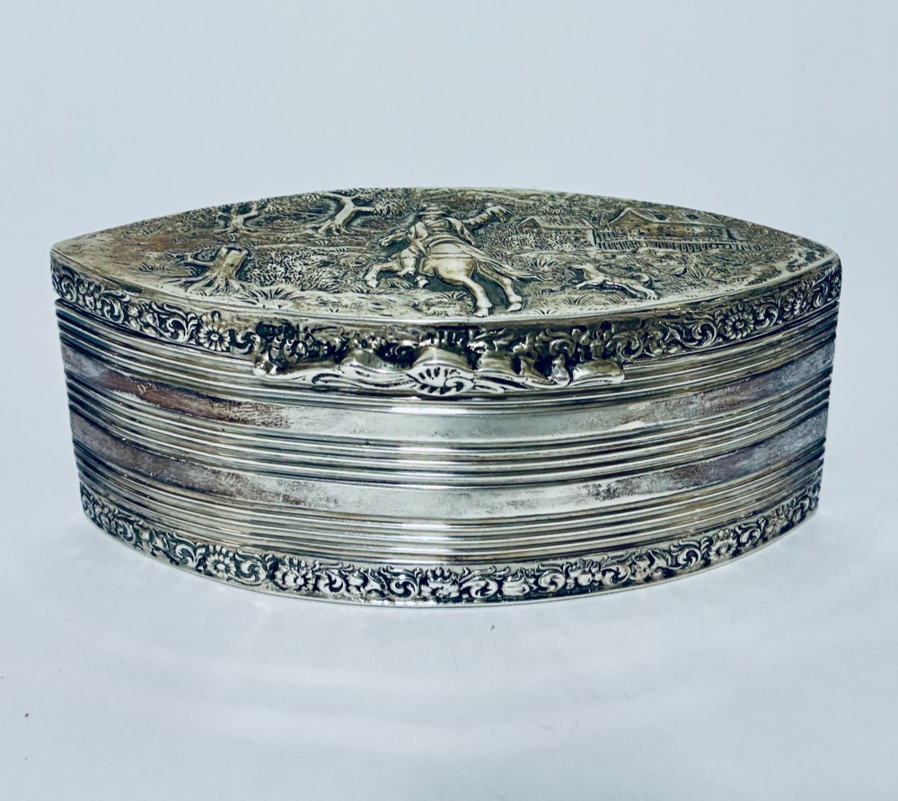 An oval silver snuff box featuring a relief equestrian scene on the hinged lid and reeded side walls with carved scroll borders.