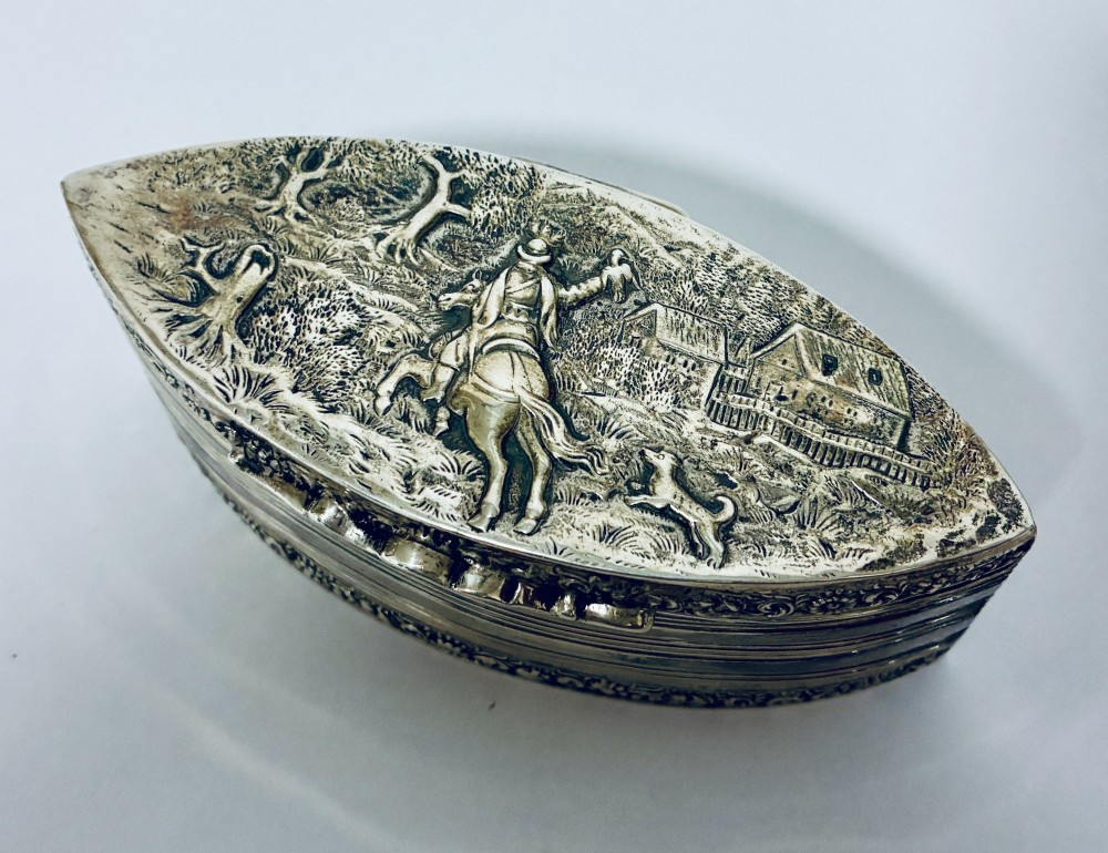An oval silver snuff box featuring a relief equestrian scene on the hinged lid and reeded side walls with carved scroll borders. View II