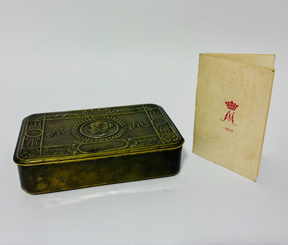 An embossed rectangular brass Princess Mary Christmas tin alongside its original cream greeting card printed with a red royal monogram.