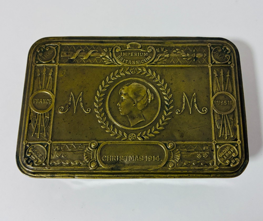 An embossed rectangular brass Princess Mary Christmas tin alongside its original cream greeting card printed with a red royal monogram. View II