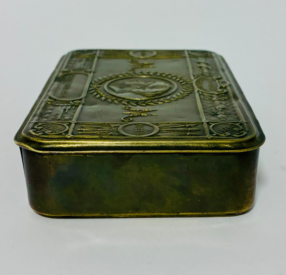 An embossed rectangular brass Princess Mary Christmas tin alongside its original cream greeting card printed with a red royal monogram. View VI