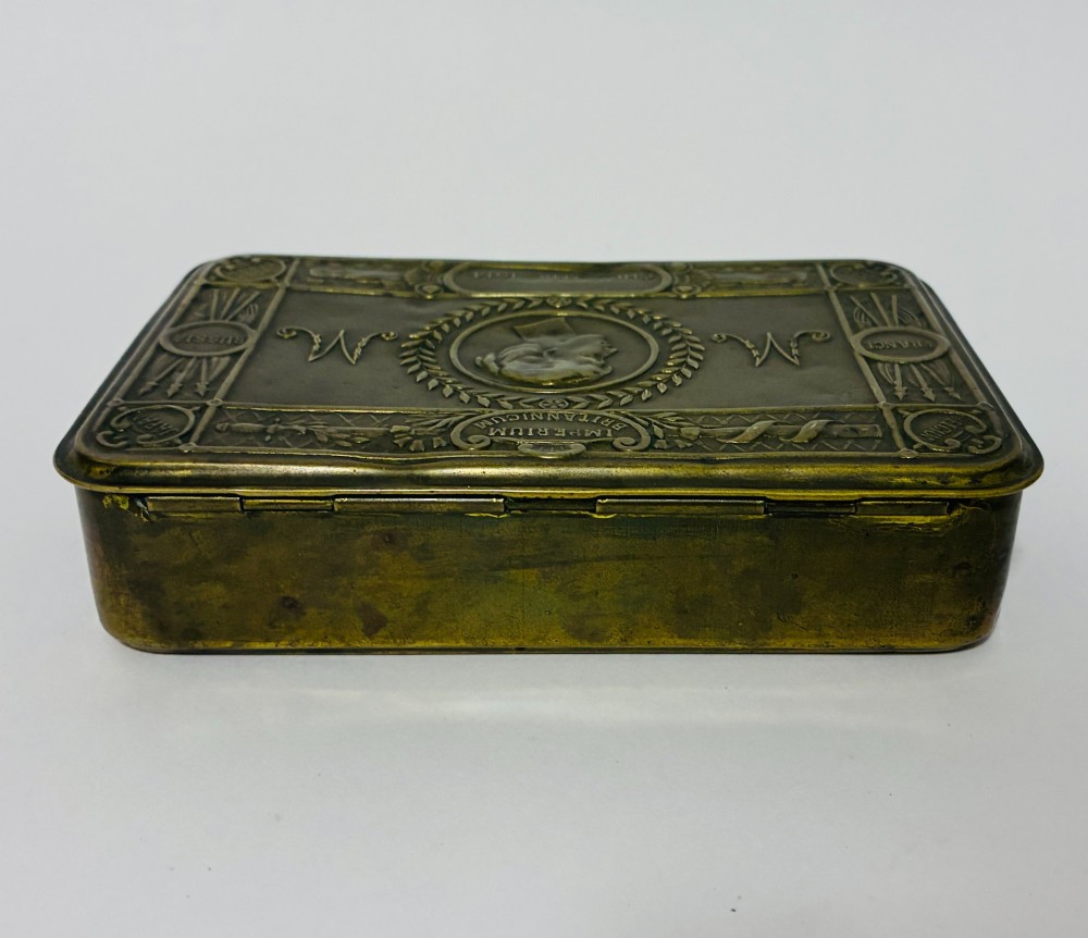 An embossed rectangular brass Princess Mary Christmas tin alongside its original cream greeting card printed with a red royal monogram. View VII