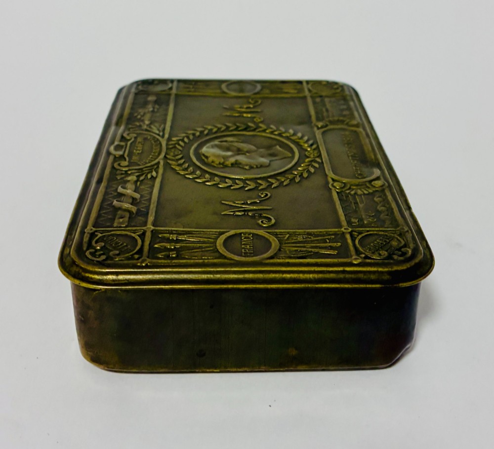 An embossed rectangular brass Princess Mary Christmas tin alongside its original cream greeting card printed with a red royal monogram. View VIII
