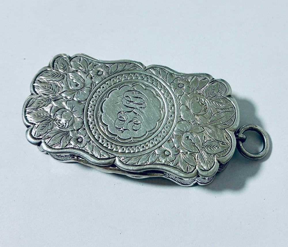 A small rectangular Victorian sterling silver vinaigrette featuring hand-engraved floral motifs, a central monogrammed cartouche, scalloped edges, and a side suspension ring.