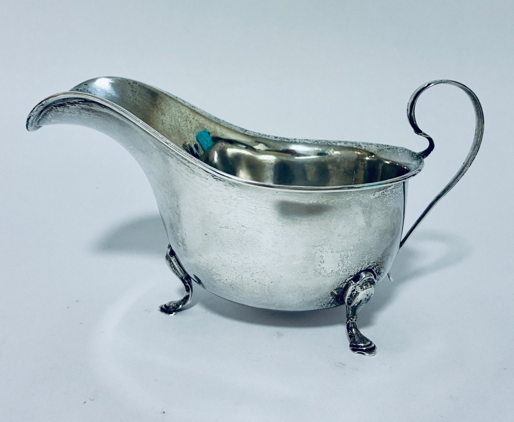 An antique silver sauce boat featuring a scrolled flying handle, flared pouring spout, and three cast pad feet.