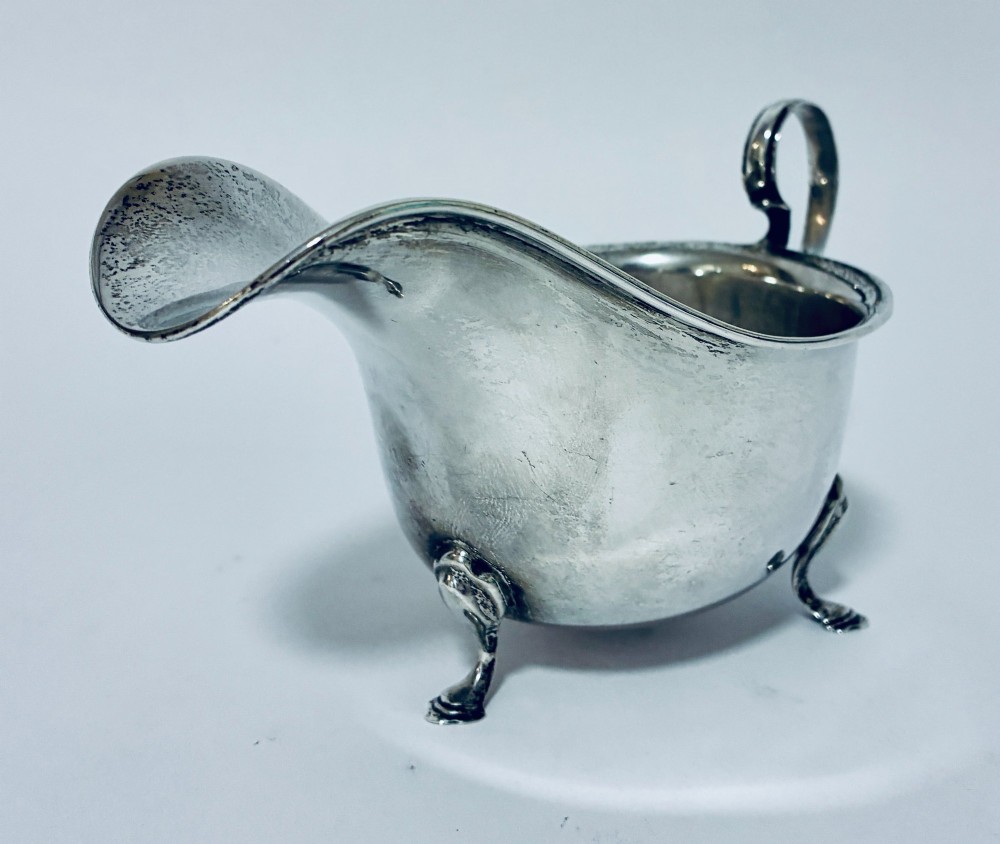 An antique silver sauce boat featuring a scrolled flying handle, flared pouring spout, and three cast pad feet. View II