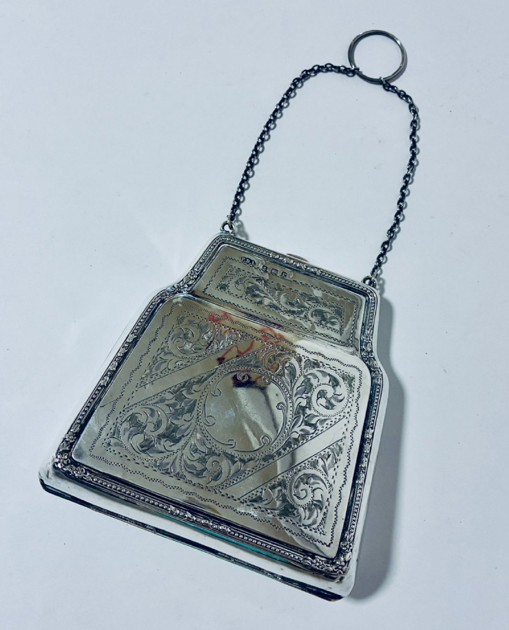 An antique sterling silver evening purse featuring engraved floral scrollwork, visible hallmarks along the upper frame, and a chain handle with a finger ring.