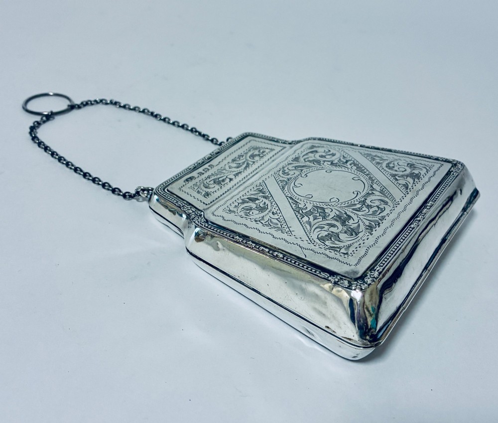 An antique sterling silver evening purse featuring engraved floral scrollwork, visible hallmarks along the upper frame, and a chain handle with a finger ring. View V