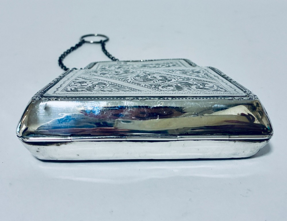 An antique sterling silver evening purse featuring engraved floral scrollwork, visible hallmarks along the upper frame, and a chain handle with a finger ring. View X